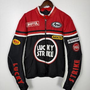 May include: A vintage motorcycle jacket in red, black, and white, featuring multiple sponsor patches. The jacket has a zip-up front, a stand-up collar, and the "LUCKY STRIKE" logo prominently displayed on the chest.