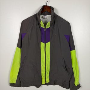 May include: A gray Puma track jacket with neon green and purple accents. The jacket features a full zipper, a high collar, and a small Puma logo on the chest. The sleeves are neon green, and the front has a vertical neon green stripe.
