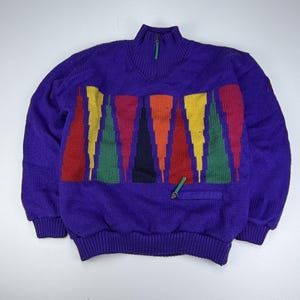 Puede incluir: Un suéter de punto morado de manga larga con cuello alto, cierre de cremallera y tirador verde. La parte delantera presenta un patrón geométrico de triángulos verticales en rojo, amarillo, verde, naranja y negro. Incluye un pequeño bolsillo frontal.