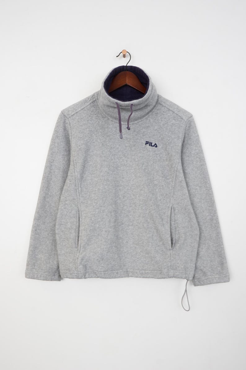 Pu&ograve; includere: Un pullover in pile grigio con collo alto foderato blu navy e coulisse. Il logo Fila &egrave; ricamato sul petto. Il pullover ha due tasche laterali ed &egrave; appeso a una gruccia di legno.