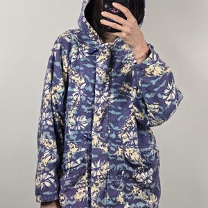 May include: A hooded fleece jacket in shades of blue, cream, and yellow, featuring a floral pattern. The jacket has a toggle closure and two front pockets. The hood is up, and the jacket appears to be worn.