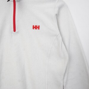 Op de afbeelding: Witte fleece trui met een rode rits en de rode letters "HH" geborduurd op de borst. De top met lange mouwen heeft een hoge kraag en een zacht, getextureerd uiterlijk. Het kledingstuk is ontworpen voor warmte en comfort.