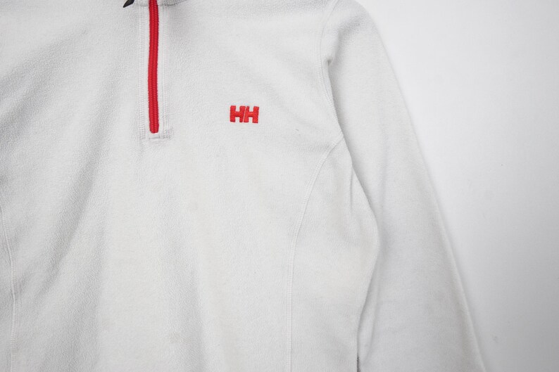 Op de afbeelding: Witte fleece trui met een rode rits en de rode letters "HH" geborduurd op de borst. De top met lange mouwen heeft een hoge kraag en een zacht, getextureerd uiterlijk. Het kledingstuk is ontworpen voor warmte en comfort.