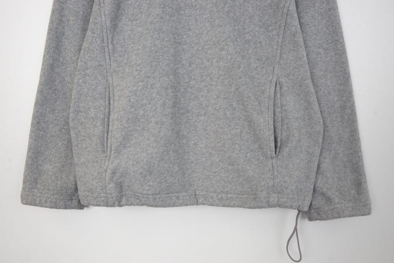 Pu&ograve; includere: Primo piano di una giacca in pile grigia. La giacca ha due tasche laterali e un cordino sull'orlo inferiore. Il tessuto &egrave; un materiale morbido e strutturato. La giacca &egrave; di colore neutro, adatta a vari outfit.