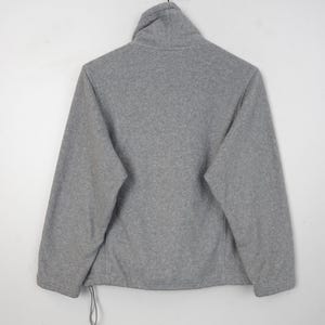 Pu&ograve; includere: Giacca in pile grigio con collo alto, maniche lunghe e coulisse sull'orlo. La giacca &egrave; appesa su uno sfondo bianco. Il materiale in pile appare morbido e caldo.