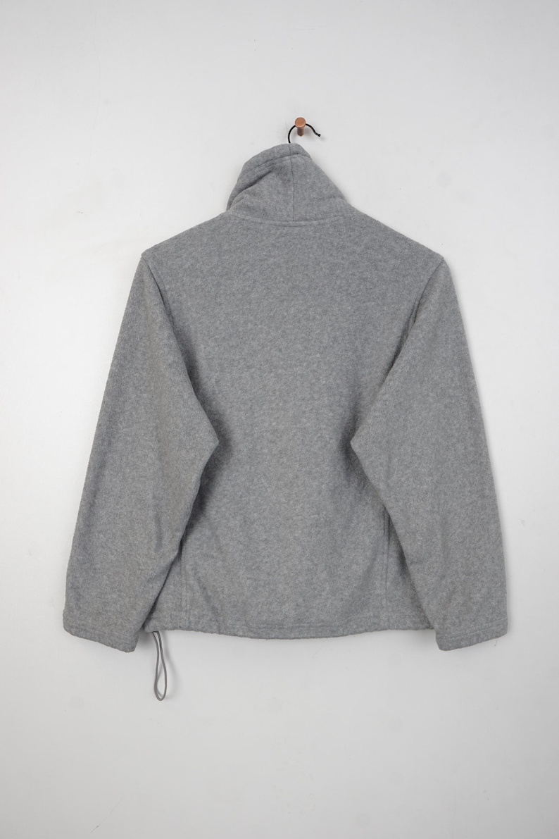 Pu&ograve; includere: Giacca in pile grigio con collo alto, maniche lunghe e coulisse sull'orlo. La giacca &egrave; appesa su uno sfondo bianco. Il materiale in pile appare morbido e caldo.