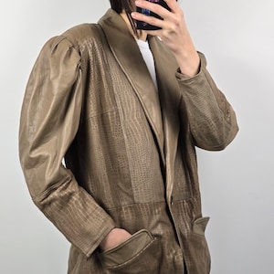 May include: A tan faux leather blazer with a crocodile skin texture. The blazer has a wide lapel, two front pockets, and padded shoulders. The sleeves are long and the person is wearing a white shirt underneath.