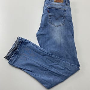 Może przedstawiać: Jasnoniebieskie jeansy z denimu o klasycznym kroju z pięcioma kieszeniami, beżową naszywką z imitacji skóry na tylnym pasku, małą czerwoną metką na tylnej kieszeni oraz lekko spranym wykończeniem z widocznymi szwami wzdłuż szwów.