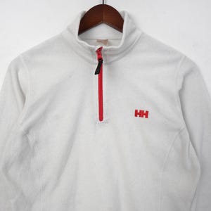 Op de afbeelding: Een witte fleece trui met een rode rits en de rode letters "HH" geborduurd op de borst. De trui met lange mouwen heeft een hoge kraag en hangt aan een houten hanger.