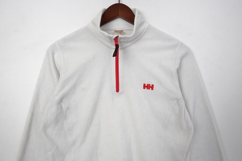 Op de afbeelding: Een witte fleece trui met een rode rits en de rode letters "HH" geborduurd op de borst. De trui met lange mouwen heeft een hoge kraag en hangt aan een houten hanger.