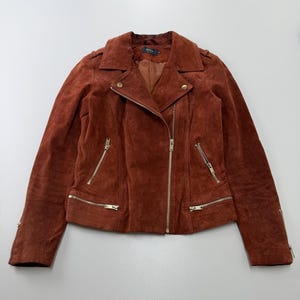 Könnte beinhalten: Eine rostfarbene Wildleder-Motorradjacke mit asymmetrischem Frontreißverschluss, breitem Reverskragen mit Druckknöpfen und mehreren silberfarbenen Reißverschlusstaschen an der Vorderseite und den Ärmeln, ausgestattet mit einem glatten Innenfutter.