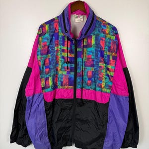May include: A vintage windbreaker jacket with a vibrant geometric pattern in shades of pink, purple, blue, and yellow. The jacket features a full zipper, a high collar, and black accents on the sleeves and lower section.