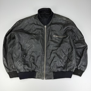 Könnte beinhalten: Eine schwarze Leder-Bomberjacke mit geripptem Kragen, Bündchen und Saum. Die Jacke verfügt über einen durchgehenden silberfarbenen Metallreißverschluss, zwei schräge Seitentaschen und eine einzelne horizontale Reißverschlusstasche auf der Brust.