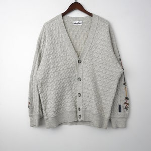 May include: A grey cable knit cardigan with a v-neck and button fastening. The cardigan has embroidered details on the sleeves and a "Gato Gato" label.