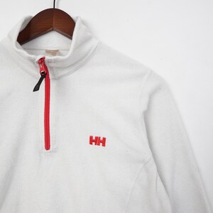 Op de afbeelding: Een witte fleece trui met een rode rits en de geborduurde letters "HH" in rood. De kraag is een col. Het kledingstuk hangt aan een houten hanger.