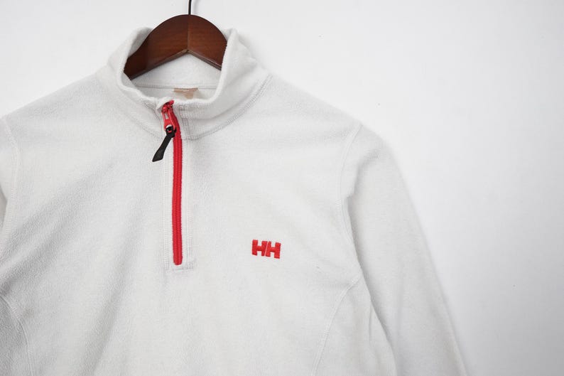 Op de afbeelding: Een witte fleece trui met een rode rits en de geborduurde letters "HH" in rood. De kraag is een col. Het kledingstuk hangt aan een houten hanger.