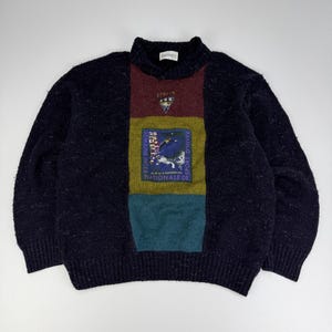 Könnte beinhalten: Ein dunkelblauer, gesprenkelter Strickpullover mit einem zentralen vertikalen Einsatz in den Farben Bordeaux, Olivgrün und Petrol. In der Mitte befindet sich ein rechteckiger Aufnäher mit dem Text "SEMAINE INTERNATIONALE SPORT D'HIVER".