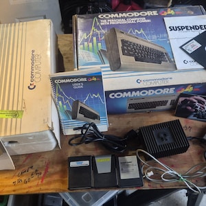 May include: A vintage Commodore 64 computer set including the original box, a 1541 floppy disk drive box, user manuals, a black floppy disk, a power supply unit, and various software booklets. The items feature the Commodore logo and branding.