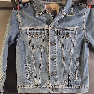 May include: A medium-wash blue denim jacket featuring a classic collar, long sleeves with buttoned cuffs, and a front button closure. The garment includes two chest pockets with buttoned flaps and two vertical side pockets, finished with visible stitching.