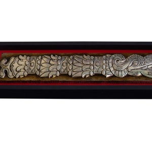 May include: A silver-colored metal relief sculpture of a stylized serpent or dragon with intricate details, framed in black with a red mat.