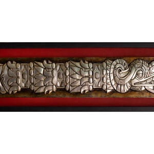May include: A silver framed artwork depicting a stylized, detailed, silver relief of a serpent with a feathered headdress. The artwork is framed in a black frame with a red mat.