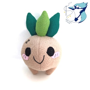 May include: A plush toy in the shape of a tan, round creature with green leaves and a light green top. It has black eyes, pink cheeks, and a stitched mouth. The toy has small tan feet. The logo for "Geeky Felt Friends" is in the upper right corner.