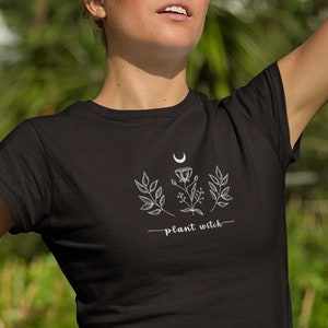 Może przedstawiać: Czarny t-shirt z białym nadrukiem przedstawiającym p&oacute;łksiężyc, liście i kwiaty. Pod nadrukiem znajduje się napis "plant witch".