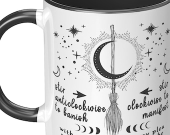 May include: White ceramic mug with a black rim and handle. The mug features a black and white illustration of a broom, a crescent moon, and stars. The text reads "stir anticlockwise to banish with each stir 1 set my plea as 1 thinketh so mote it be" and "stir clockwise to manifest".