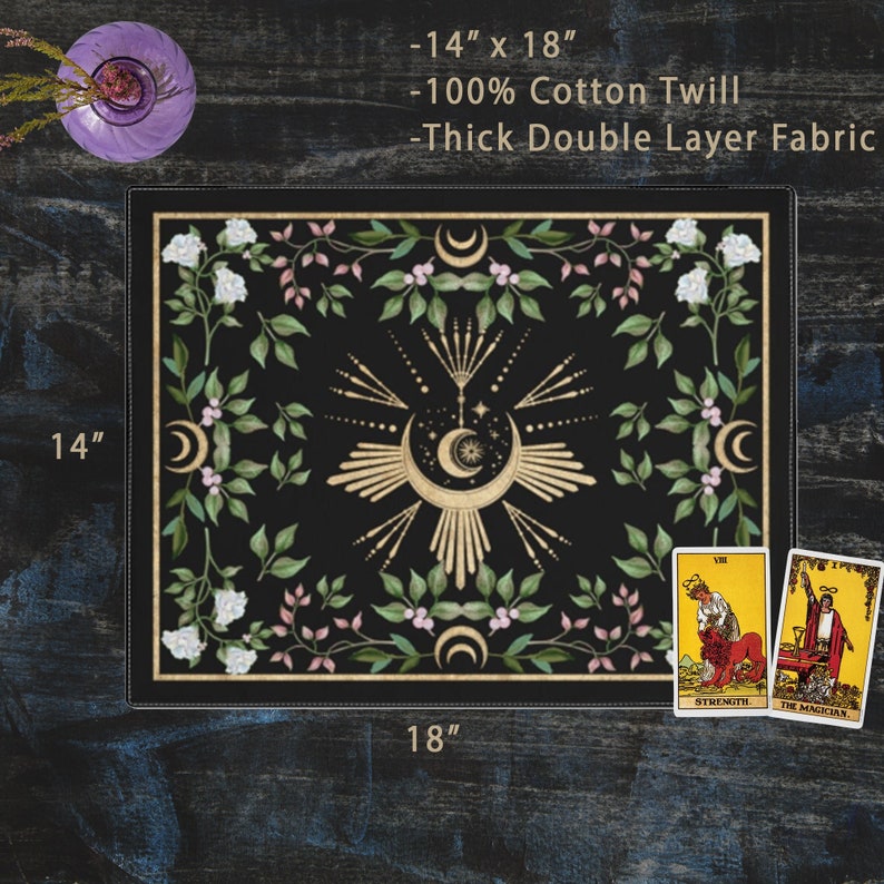 Op de afbeelding: Een zwarte tarotdoek met een gouden maan- en sterrendesign omringd door witte bloemen en groene bladeren. De doek meet 35,56 cm bij 45,72 cm en is gemaakt van 100% katoenen keper. De doek is dik en dubbel gelaagd. Twee tarotkaarten worden getoond in de rechterbenedenhoek: Kracht en De Tovenaar.