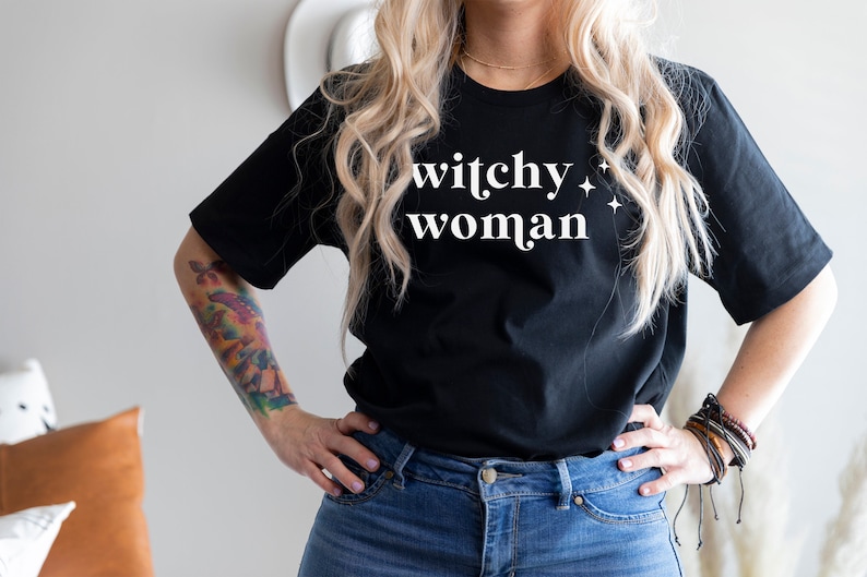 Pu&ograve; includere: Una maglietta nera con il testo "witchy woman" in lettere bianche e piccole stelle bianche.