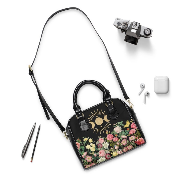 May include: Black leather satchel handbag with a gold sun and moon design and floral print. The bag has a top handle and a long adjustable shoulder strap.