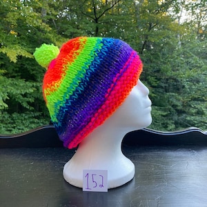 May include: A rainbow knit beanie with a green pom pom. The beanie is on a white mannequin head. The number 152 is written on a white tag.