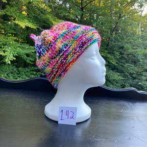 May include: A colourful knitted hat with a white background and stripes of pink, purple, green, yellow, and orange. The hat is on a white mannequin head with the number 142 written on a white tag.