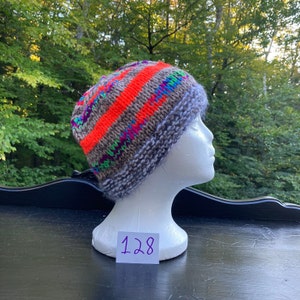 May include: A grey knitted hat with a wide orange stripe and colourful accents. The hat is on a white mannequin head with a tag that says "128".