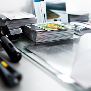 May include: Stacks of printed cards with colorful images, possibly postcards or art prints, are arranged on a reflective surface. A black and silver tool and a black utility knife are in the foreground. The cards feature landscape and floral designs.