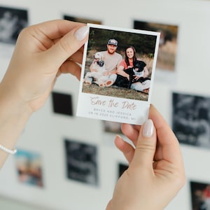 May include: A white polaroid-style photo held by a person. The photo features a couple with two dogs and the text "Save the Date." The date and location are also printed on the photo. Other photos are visible in the background.