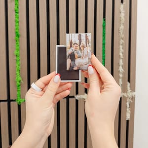 May include: A rectangular photo magnet with a picture of a couple and a dog. The magnet is held in front of a wooden panel with vertical brown stripes. The person holding the magnet has red nail polish.