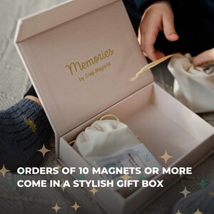 a person's hands opening a gift box with a white interior and gold text that reads "Memories by Snap Magnets".