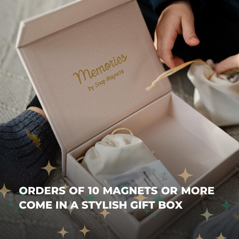 a person's hands opening a gift box with a white interior and gold text that reads "Memories by Snap Magnets".