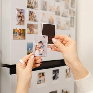 May include: A collection of rectangular photo magnets, some attached to a white refrigerator. One magnet is being held up, showing a family portrait. The magnets are a mix of family photos and portraits.