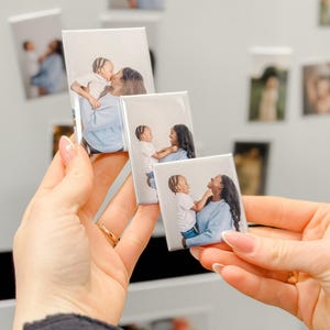 May include: Rectangular photo magnets featuring a mother and child. The magnets display a series of images of a mother and child interacting. The magnets are held in a person's hand, with other magnets visible in the background.