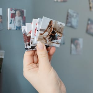 May include: A fan of small, square, glossy photo magnets featuring various printed images, including a person and a child on a wooden floor, a building, and other scenes, held together in a stack to display the individual photographic prints.