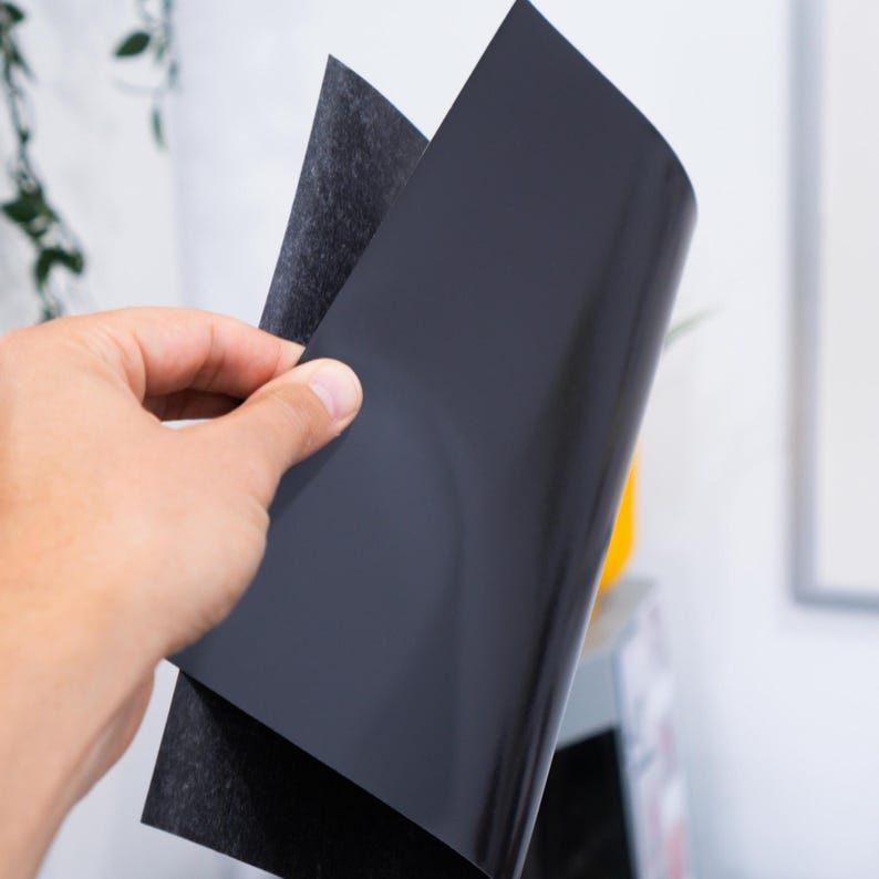 May include: A hand holding two sheets of black magnetic material. One sheet has a smooth, glossy finish, while the other has a textured, felt-like surface. The sheets are slightly curved, suggesting flexibility.