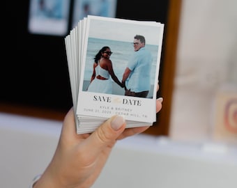 Puede incluir: Una pila de tarjetas blancas de Save the Date con una pareja tomados de la mano en una playa. Las tarjetas tienen el texto "SAVE DATE" y los nombres "Kyle & Britney" con la fecha y el lugar de la boda.