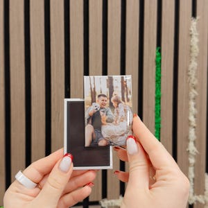 A person's hands holding a small photo frame with a picture of a couple inside, against a wooden background with vertical black and white stripes.