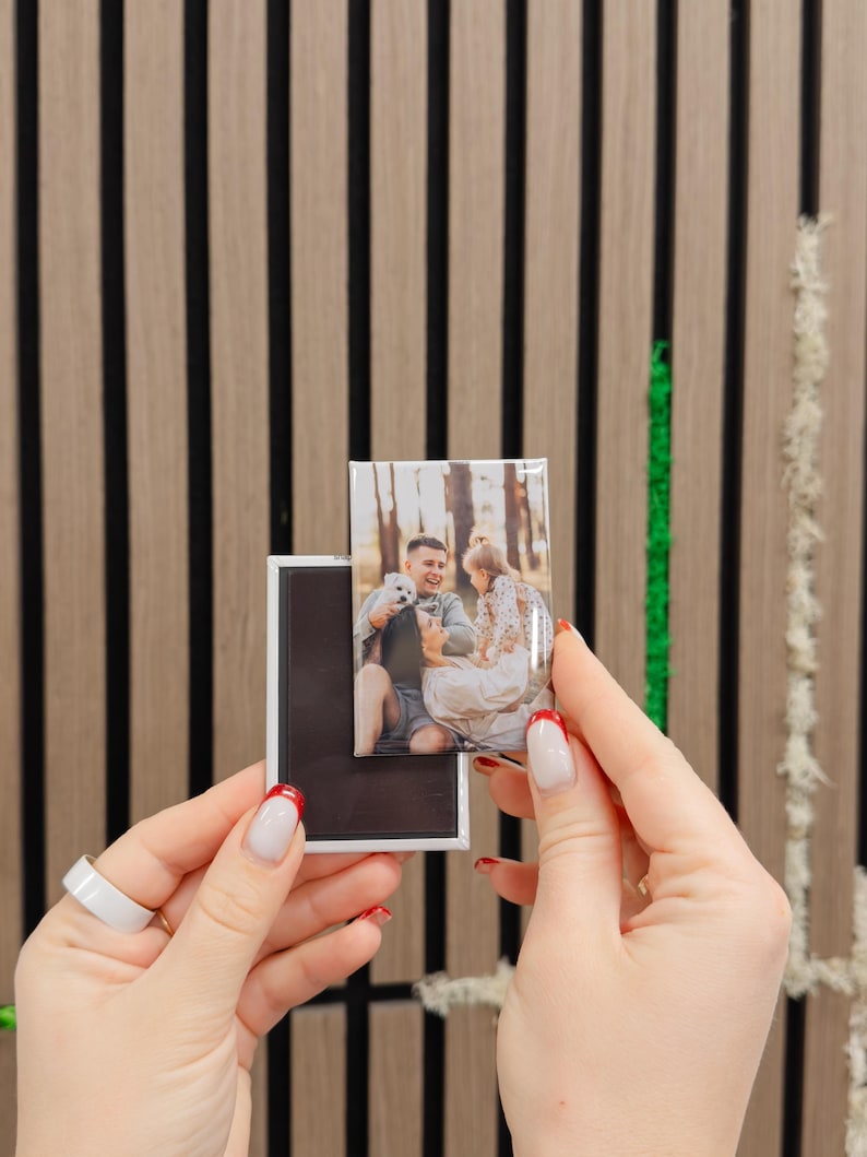 A person's hands holding a small photo frame with a picture of a couple inside, against a wooden background with vertical black and white stripes.