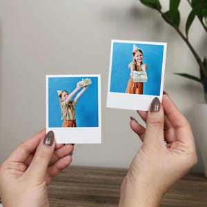 May include: Two square instant film prints featuring a child with red hair wearing a gold sequined top and orange trousers against a blue background. In one print, the child holds a gift box overhead; in the other, the child holds the gift forward.