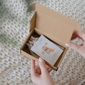 A person's hands are holding an open cardboard box containing a small white card with an illustration of a hand holding a smartphone.