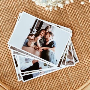 May include: A stack of rectangular photographic prints featuring a colour image of a family of three embracing. The top photograph shows a parent in a black and white striped shirt and another parent in a dark shirt holding a small child.