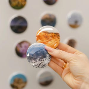 May include: Two circular pin-back buttons featuring photographic prints. One button displays a vibrant orange desert landscape, while the other shows a blue and white snowy mountain range. Both buttons have a glossy finish and rounded edges.